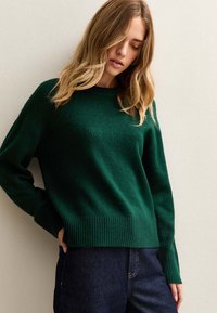 Groene gebreide trui met een ronde hals, geribbelde zoom en lange mouwen, gecombineerd met donkere denim. Glad textuur en een casual pasvorm.