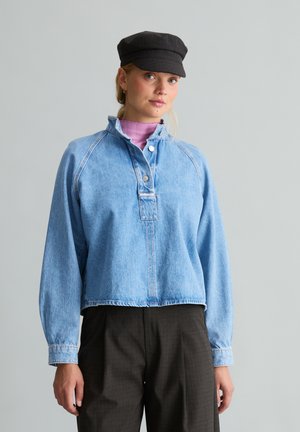 Junge Frau trägt einen hellblauen Denim-Pullover, schwarze Mütze, lila Rollkragen und dunkle Hose und steht vor einem einfarbigen grauen Hintergrund.