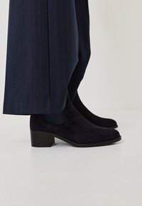 Botines de ante azul marino oscuros con un tacón bajo y paneles laterales elásticos, combinados con pantalones de rayas azul marino. Superficie texturizada visible.