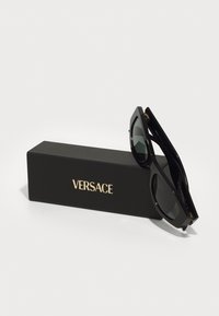 Gafas de sol negras de Versace expuestas sobre una caja negra con letras doradas. Presentan montura angular y lentes oscuros con un acabado brillante.