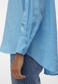 Camicia azzurra chiara, con una texture, maniche lunghe, con due bottoni ai polsini e un orlo irregolare, abbinata a jeans di denim.