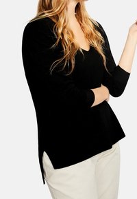 Haut noir à manches longues avec un col en V, fentes latérales et coupe décontractée, associé à un pantalon beige clair. Texture douce, design minimal.