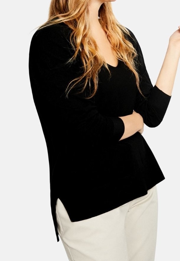 Haut noir à manches longues avec un col en V, fentes latérales et coupe décontractée, associé à un pantalon beige clair. Texture douce, design minimal.