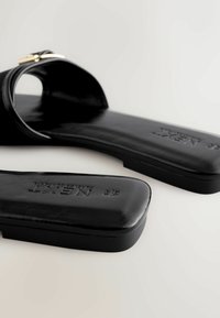 Sandales plates noires avec bout carré, large bride avec détail boucle dorée, et logo de la marque embossé sur la semelle, sur fond blanc.