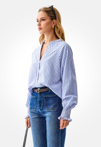 Blauw-wit gestreepte blouse met knopen, met ballonmouwen en geplooide manchetten, gecombineerd met een high-waisted denim jeans en een bruine riem.
