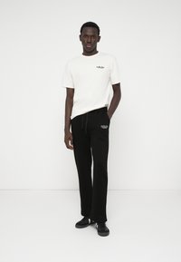 Λευκό βαμβακερό t-shirt με μικρό γραφικό στο στήθος, συνδυασμένο με μαύρες παντελόνες jogger με κεντημένο λογότυπο, φορεμένες με μαύρα αθλητικά παπούτσια.