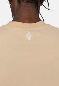 Béžové bavlněné tričko s jednoduchým designem, které má na horní straně zad malé bílé geometrické vyšívané logo.