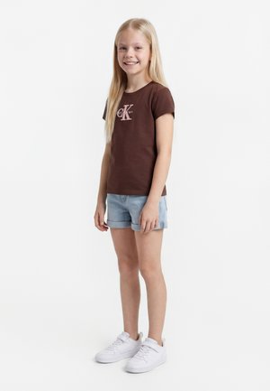 Ung pige står iført en brun Calvin Klein t-shirt, lyseblå op-rullede denimshorts og hvide sneakers, smilende til kameraet.
