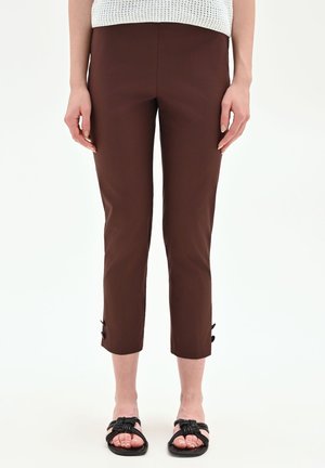 Donna che indossa pantaloni marroni corti con dettagli a bottoni vicino alle caviglie, abbinati a sandali neri intrecciati e top bianco senza maniche in maglia.