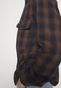 Brun och marinblå rutig flanellskjorta med knapplist, sidficka och mjukt material, bärs över grå jeans, vilket visar en sidoprofil.