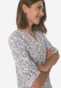 Blusa a maniche lunghe con scollo a V, caratterizzata da un motivo floreale in blu, viola e verde su sfondo bianco, realizzata in tessuto leggero.