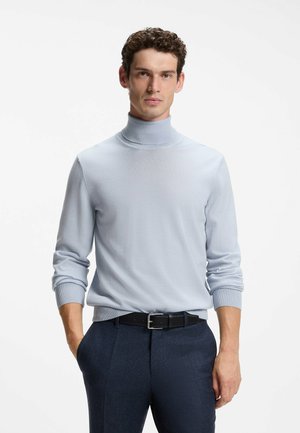 Giovane uomo con capelli ricci che indossa un maglione dolcevita azzurro chiaro e pantaloni blu scuro, in piedi davanti a uno sfondo bianco semplice.