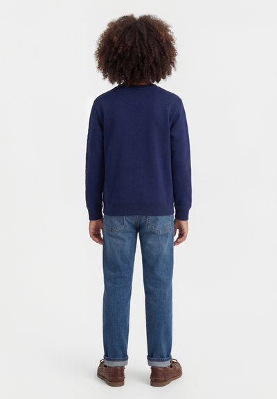 Παιδί με σγουρά μαλλιά στέκεται πλαγίως, φοράει μπλε ναυτικό φούτερ, μπλε τζιν με γυρισμένα μπατζάκια και καφέ παπούτσια σε λευκό φόντο.