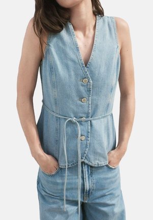 Kvinde iført ærmeløs lyseblå cowboyvest med knapper og et bundet bælte, hænder i lommerne, kombineret med matchende cowboybukser.