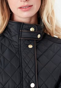 Black quilted jacket with a high collar, featuring gold hardware accents and brown piping along the edges. Textured surface with diamond patterns.