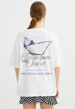 Mujer con coleta castaña que lleva una camiseta blanca oversize con un dibujo azul y negro de un barco de papel y texto sobre la vela y Saint-Tropez.