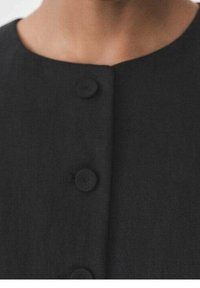 Černý oděv s kulatým výstřihem, strukturovanou tkaninou a dvěma velkými knoflíky uprostřed. Textura látky vypadá hladce a matně.