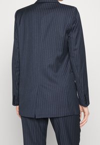 Vue de dos d'une personne portant une veste de costume bleu marine à fines rayures et un pantalon assorti, debout devant un fond uni.