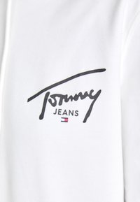 Tela blanca con el logotipo "Tommy Jeans" en negro, con una textura suave y una fuente cursiva fluida. Pequeño acento tricolor debajo del texto.