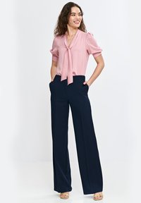 Jasnoróżowa bluzka z wiązaniem, krótkie bufiaste rękawy, w parze z granatowymi, szerokimi spodniami o wysokim stanie. Modelka stoi z rękami w kieszeniach.