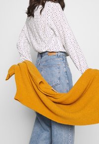 Camicia bianca a maniche lunghe con motivo a pois neri e dorati, abbinata a jeans azzurri a vita alta e un maglione lavorato di colore giallo senape.