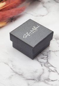 Svart, texturerad smyckeslåda med lock, med vit branding på toppen. Basen är fyrkantig med rena kanter, placerad på en marmoryta.