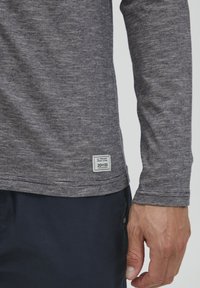 Langærmet grå T-shirt med en meleret tekstur, der har et mærke i kanten. Båret sammen med mørke marineblå bukser.