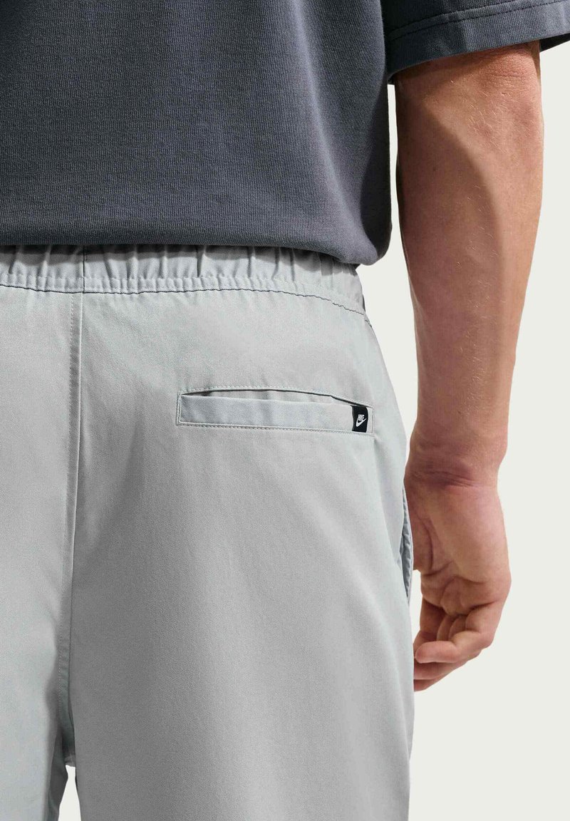 Primo piano di una persona che indossa pantaloni grigio chiaro con vita elastica e tasca posteriore con una piccola etichetta con il logo Nike, e una maglietta grigio scuro.