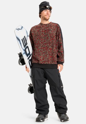 Snowboarder portant un pull à motifs rouge et noir, un pantalon ample noir et des bottes noires, tenant un snowboard blanc avec des accents bleus.
