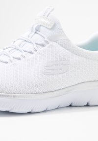 Scarpa sportiva bianca con tomaia in mesh testurizzata, lacci elasticizzati e suola ammortizzata. Presenta un logo sul lato.