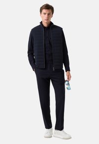 Gilet matelassé marine sur une chemise sombre, assorti de pantalons. Le modèle tient des lunettes de soleil bleues ; il porte des baskets blanches. Design moderne et minimal.