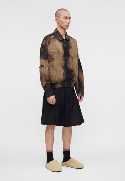 Άτομο που φοράει μια φθαρμένη καφέ και μαύρη ζακέτα, μαύρη πλισέ φούστα, μαύρες κάλτσες και μπεζ χνουδωτές παντόφλες, στέκεται μπροστά σε απλό φόντο.