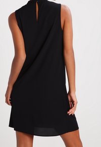 Robe noire sans manches avec un col montant et une ouverture en forme de clé au dos, présentant un design fluide et une texture lisse.