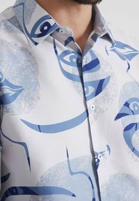 Chemise blanche avec un œil abstrait bleu et des motifs courbés, dotée d'un col écarté et d'une fermeture à boutons. Tissu lisse et léger.