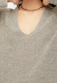 Beige gebreide trui met een V-halsontwerp, kenmerkend voor een zachte textuur en minimale ribbels. Gecombineerd met een gouden ketting.