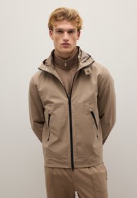 Giacca impermeabile beige con cappuccio, dotata di tasche con zip e zip anteriore a tutta lunghezza. Texture liscia con una finitura leggermente lucida.
