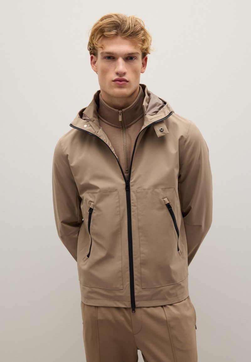 Giacca impermeabile beige con cappuccio, dotata di tasche con zip e zip anteriore a tutta lunghezza. Texture liscia con una finitura leggermente lucida.