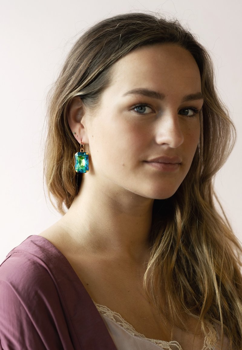 Gold-tone earrings feature a large rectangular iridescent gemstone in green and blue, suspended from a thin hook. Model wears a mauve top.