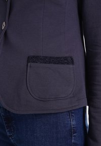 Námořnický dlouhý rukáv s kulatým lemem a texturovanou kapsou, zdobený třpytivým okrajem. Vyrobeno z měkké, hladké látky.