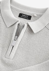 Polo en maille gris clair avec une fermeture éclair argentée et une étiquette noire "NEXT Made in Bangladesh" au col.