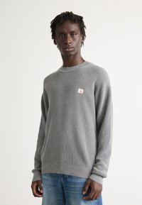 Junger Mann in einem grauen, strukturierten Strickpullover mit einem kleinen rot-weißen Logo-Patch, kombiniert mit blauen Jeans, stehend vor einem weißen Hintergrund.