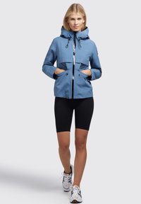Light blue waterproof jacket with a hood, two front pockets, and a zip closure, worn over black bike shorts and paired with white sneakers.