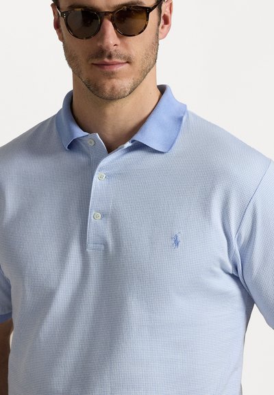 Cămașă polo de culoare albastru deschis, cu un model texturat, trei butoane și un mic logo brodat pe partea stângă a pieptului. Mâneci scurte, guler în nuanță contrastantă.