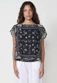 Haut noir à manches courtes avec motif paisley blanc et bord décoratif. Coupe ample avec ourlet légèrement arrondi et tissu doux et léger.