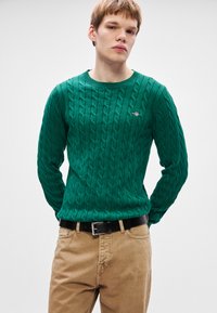 Πράσινο πλεκτό πουλόβερ με σχέδιο καλωδίων, στρογγυλή λαιμόκοψη και κεντητό λογότυπο. Συνδυάζεται με ανοιχτό καφέ παντελόνι και μαύρη ζώνη.