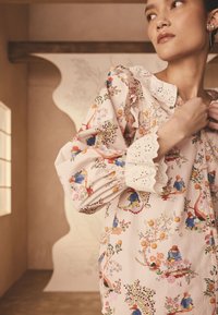 Bluzka w kwiatowy wzór z miękkiej różowej tkaniny, z bufkami na rękawach, falbankowym kołnierzykiem oraz misterną koronką na mankietach. Zapięcie na guziki.