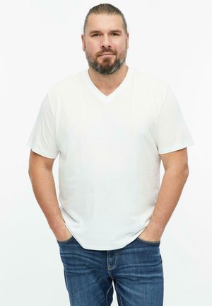 Man with beard and tied-back hair wearing a white v-neck t-shirt and blue jeans, standing with hands in pockets against a white background.