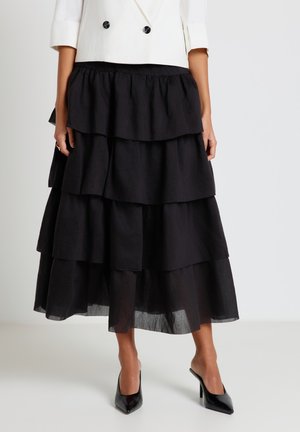 Femme portant une jupe midi noire à volants avec un blazer blanc croisé court et des escarpins noirs à bout pointu.