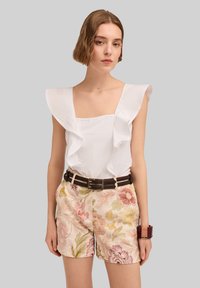 Weißes Top mit Rüschendetail, quadratischer Ausschnitt; kombiniert mit floral gemusterten Shorts in sanften Rosa-, Gelb- und Grüntönen sowie einem dunklen Gürtel mit Metallschnalle.
