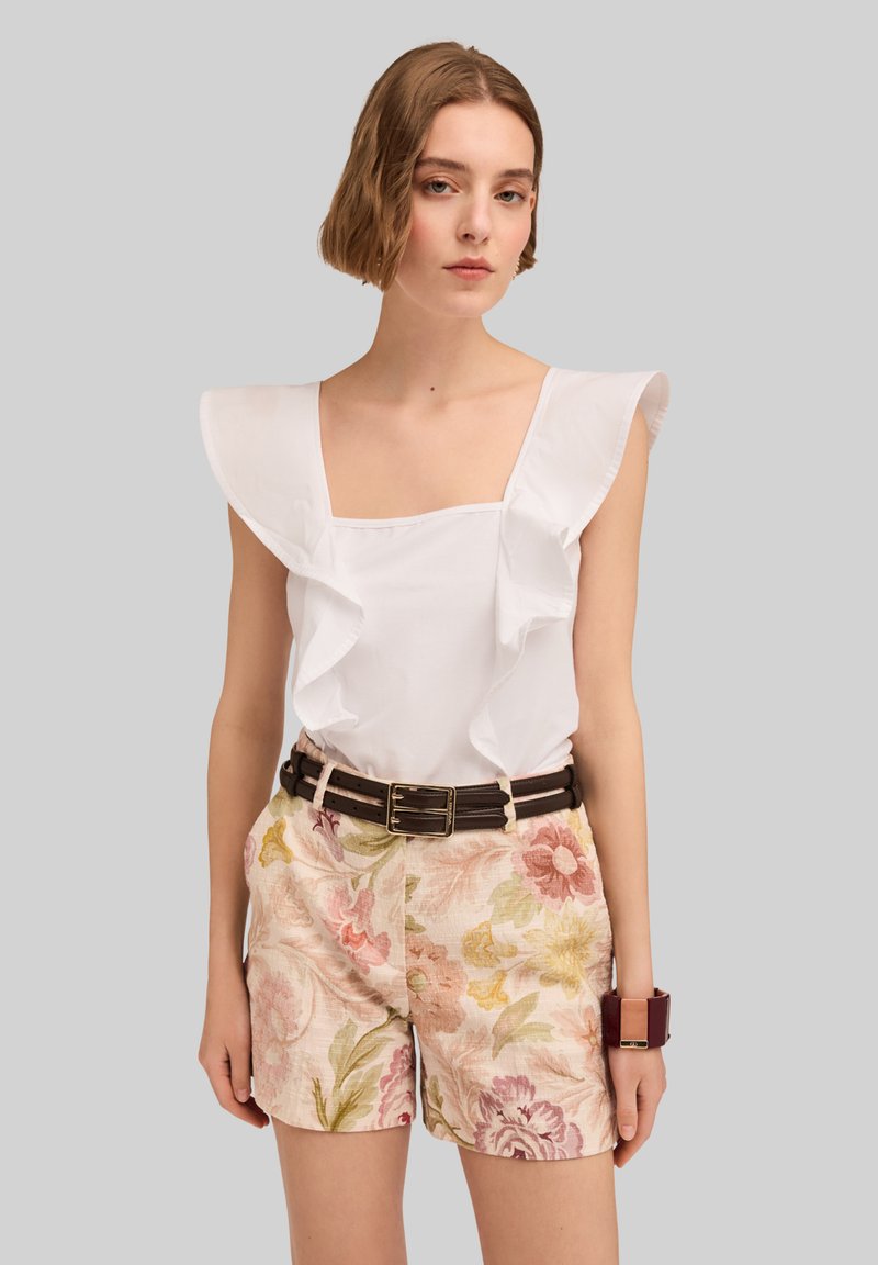 Top bianco con rouches davanti, scollatura quadrata; abbinato a pantaloni corti floreali in rosa pallido, giallo e verde e una cintura scura con fibbia in metallo.